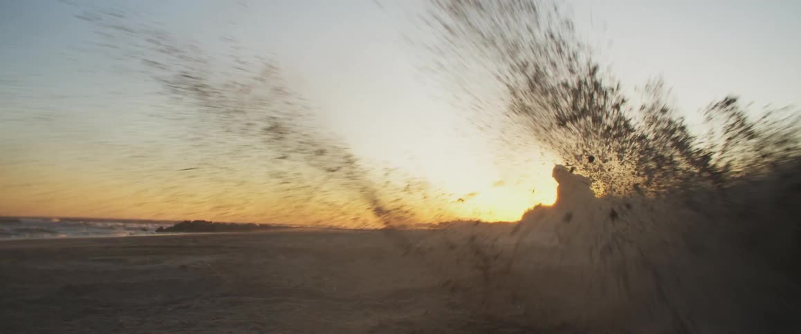 A cyclist is riding a motorcycle on a beach and doing a donut kicking up sand and dirt into the air