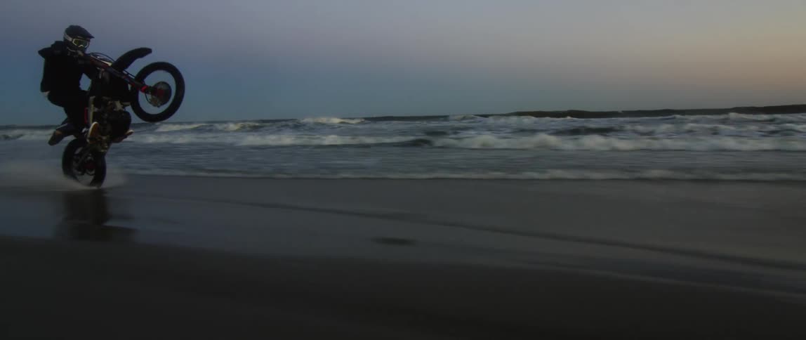Motorcycle rider doing wheelies on the shoreline at dusk