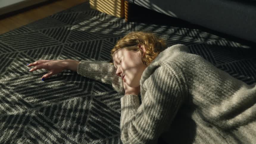 Woman with wavy hair and gray sweater lies on gray rug next to couch in living room at home