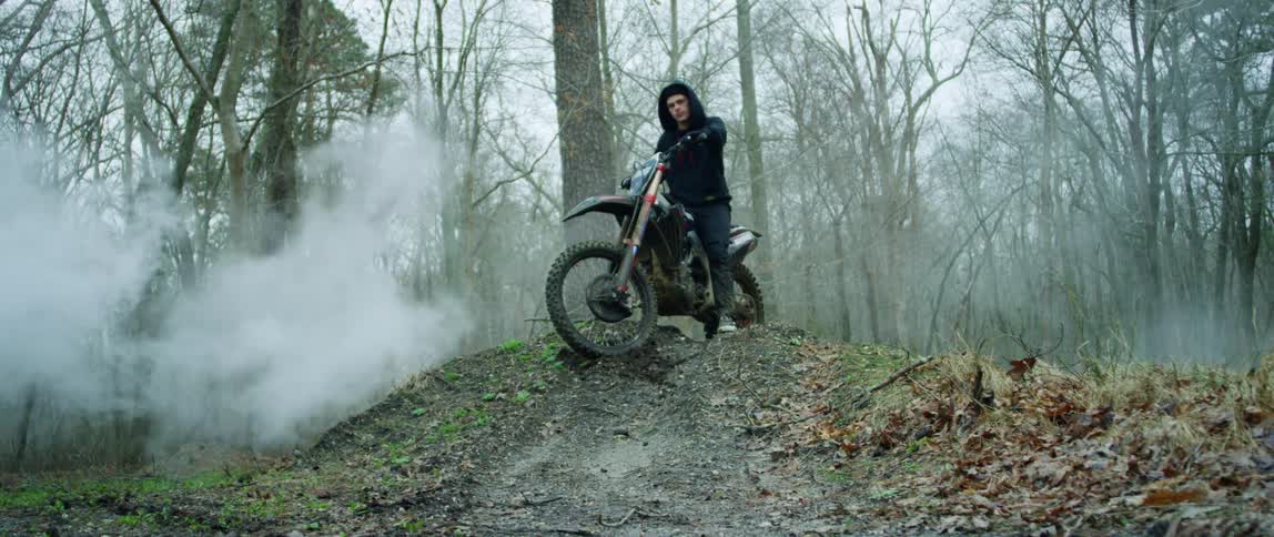 Young intense man standing over a parked motorcycle surrounded by white smoke in the forest