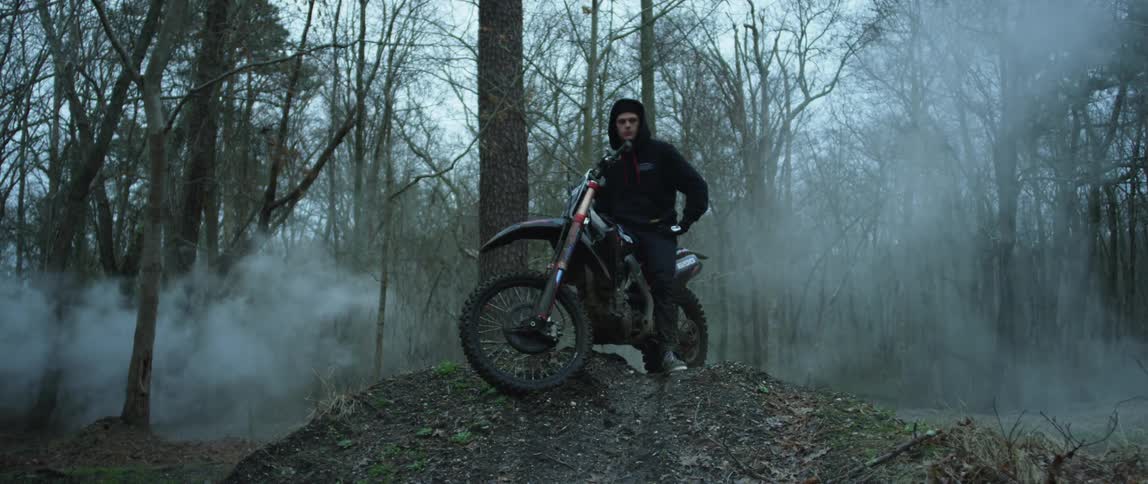Young intense man standing over a parked motorcycle surrounded by white smoke in the forest