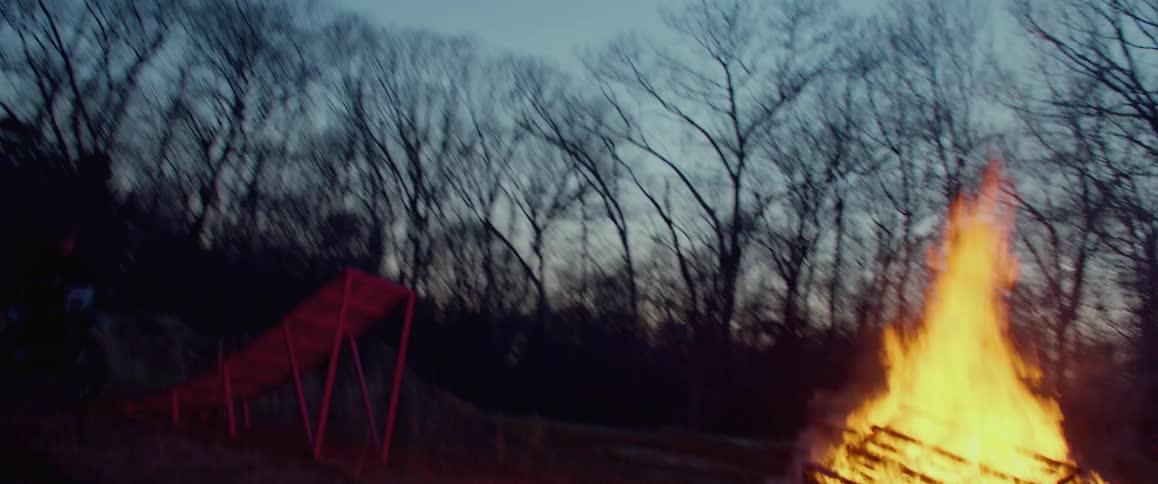 A motorcyclist rides a motorcycle up a wooden ramp to jump over a bonfire in a forest at sunset
