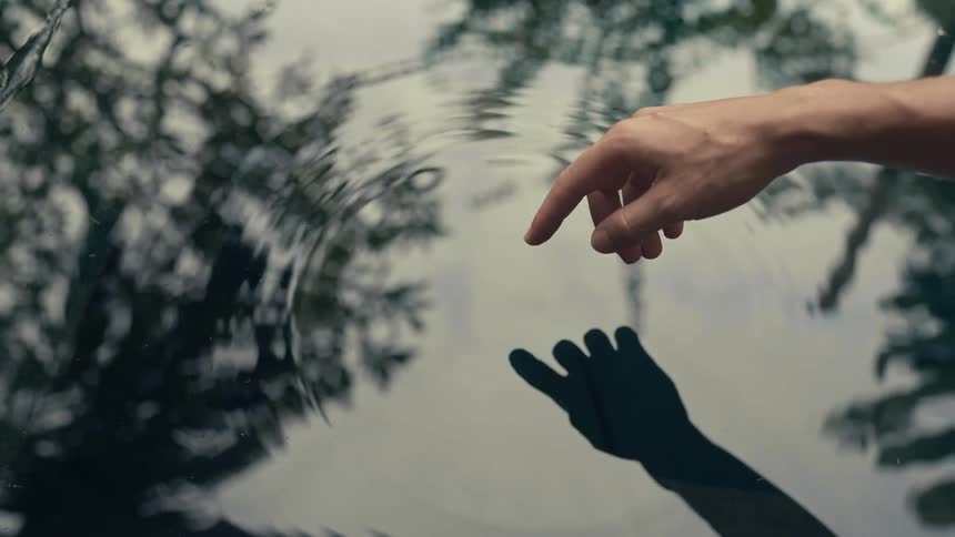 Hand touches the calm surface of a pond with a finger creating an outwards ripple