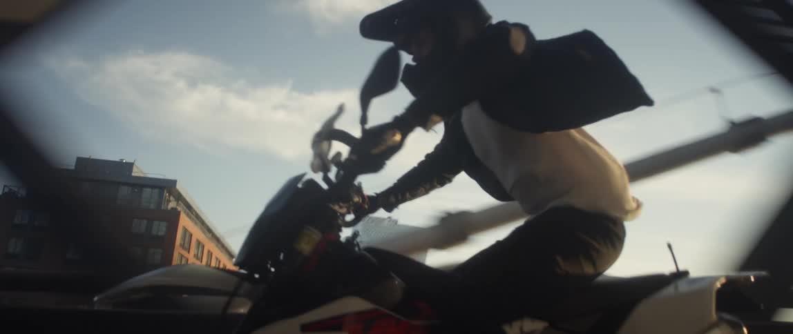 A man driving his motorcycle across a bridge in a city stands up and pops a wheelie
