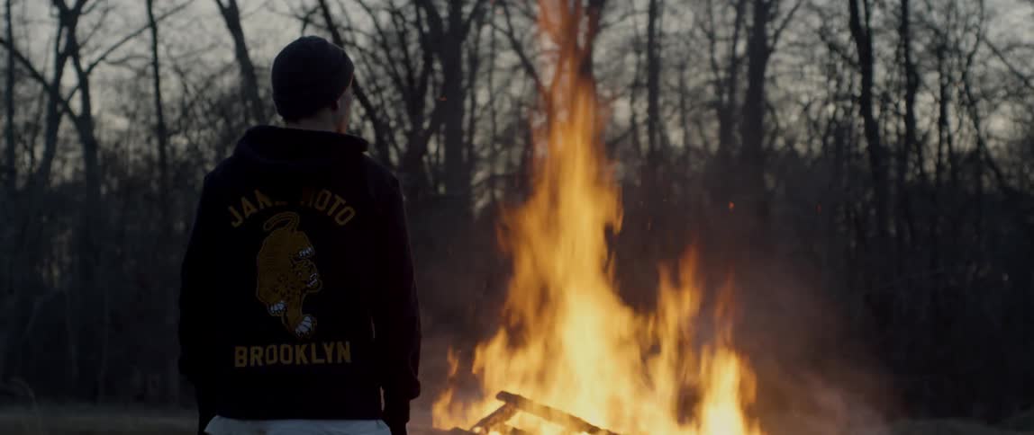 Man stands watching wood frame burn on bonfire in middle of field near leafless trees