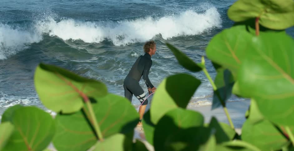 A surfer steps off his surfboard and lifts it to carry it across a beach