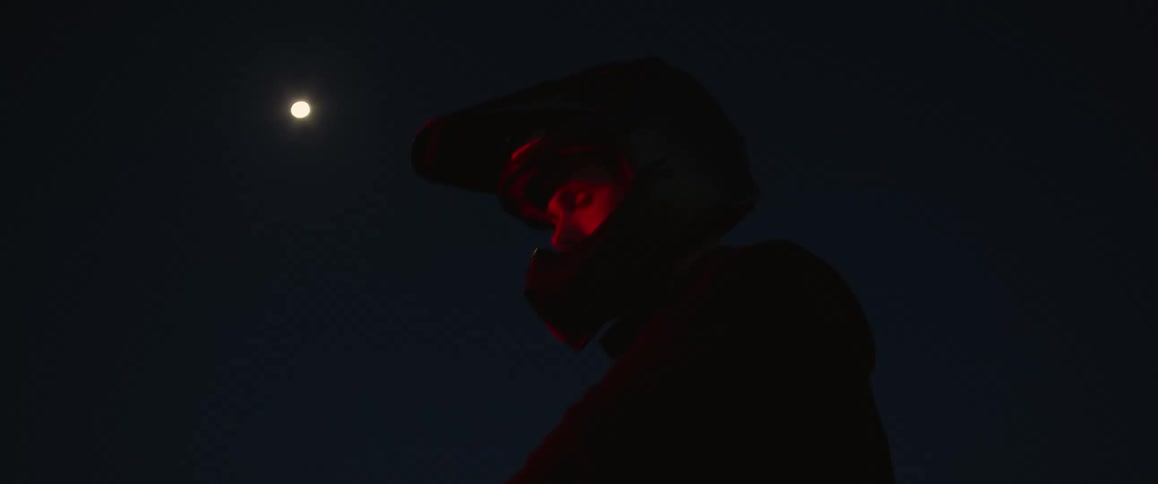 A motorcycle rider wearing a helmet is stopped and looks around with a full moon in the sky above him