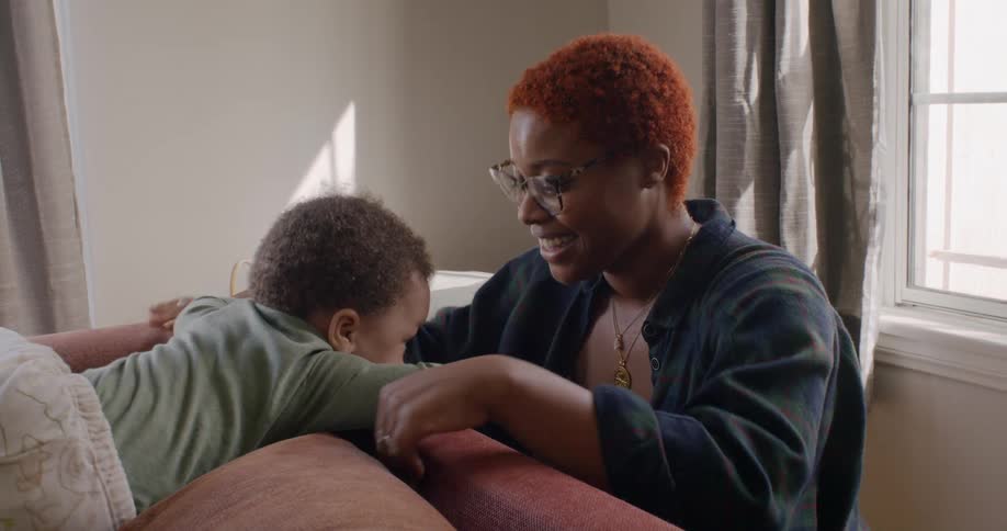 A baby stands up on a couch as his mom leans over the edge and kisses him on the head