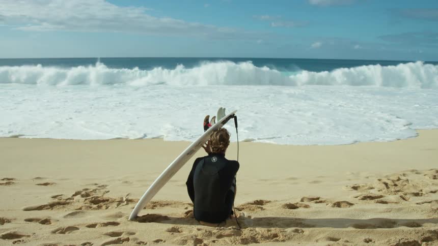 A man wearing a wet suit is sitting on a beach with his surfboard watching the waves roll in