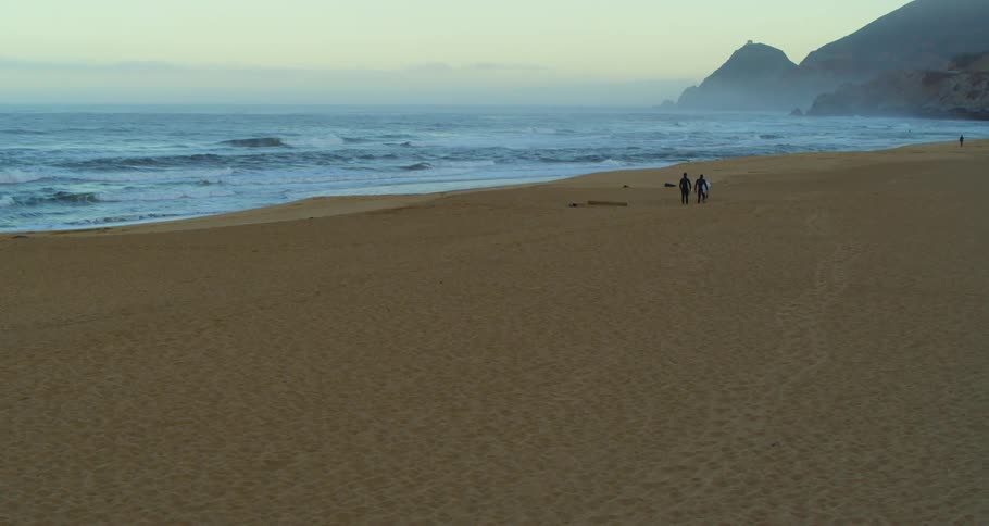 A pair of surfers walk together across a beach while carrying their surfboards on a foggy day