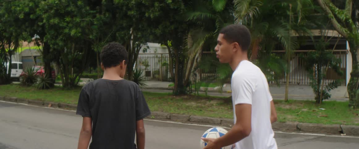 A teenager throws a soccer ball in the air as he and a friend walk through a neighborhood