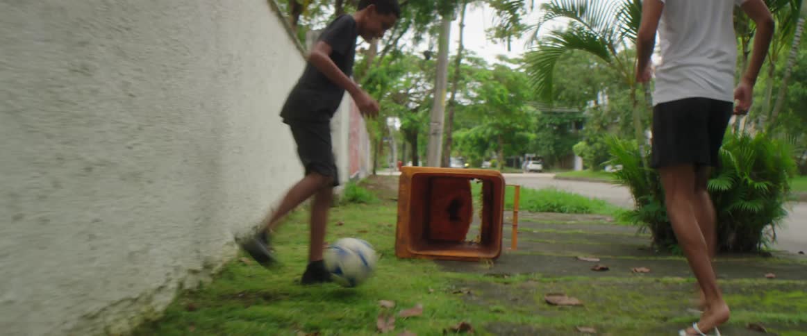 A pair of boys playing together kick a soccer ball into a tipped over trash bin outdoors during the day