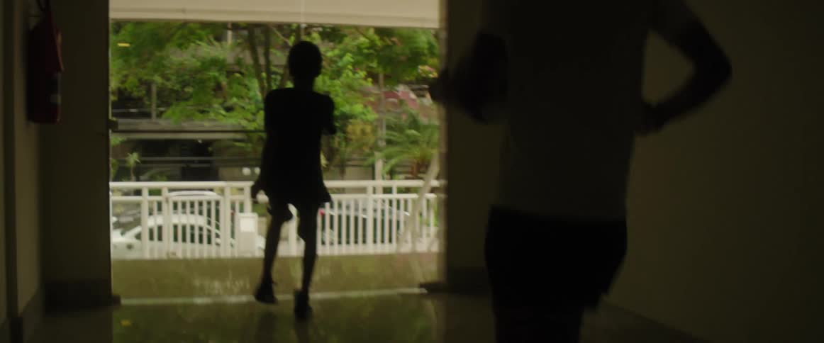 A boy excitedly walks to the exit of a building while a young man carrying a soccer ball follows behind