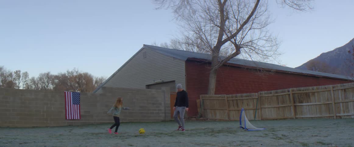 A little girl and her dad play soccer in a fenced yard