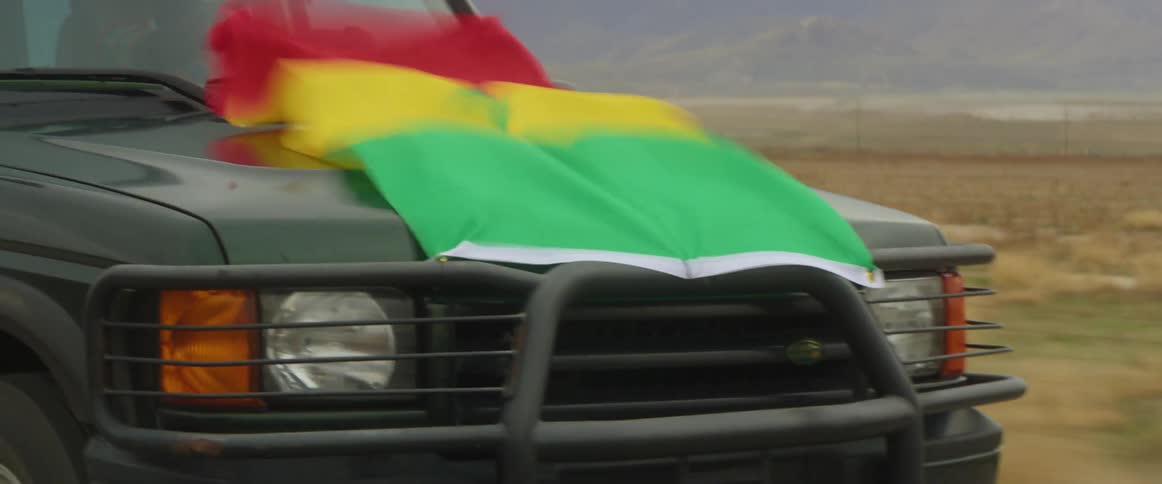A truck is traveling with an ethiopian flag on the hood and a black man sitting outside the passengers side window with his arms up in the air