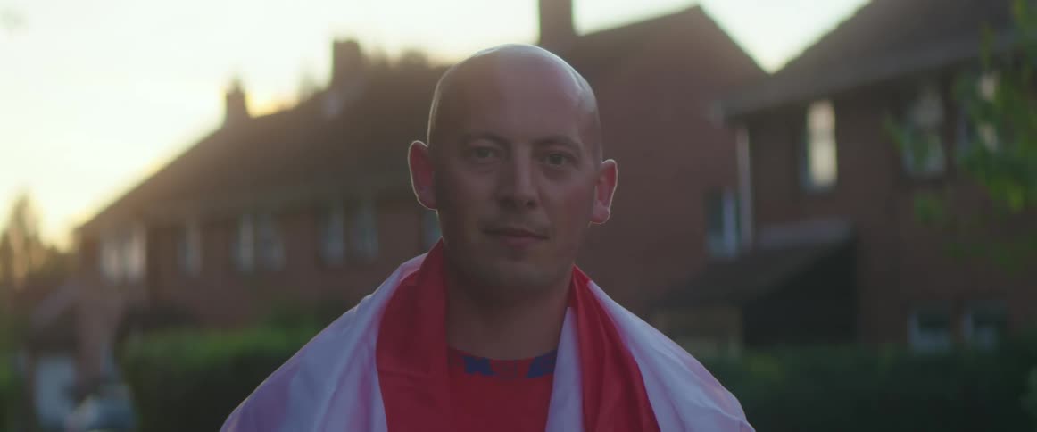 A bald man standing in a neighborhood with white and red fabric draped over his shoulders