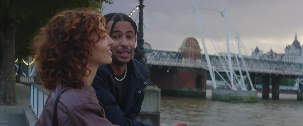 A man and woman lean on a railing on the shore and talk and laugh together as pedestrians walk past