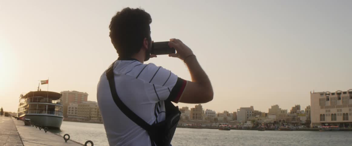 A man is sitting on a dock in a citys waterway in the daytime and using a smartphone to take a photo of buildings on the other side of the canal
