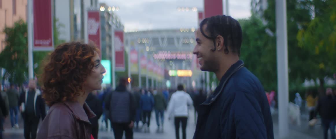 A man and woman stand on a busy street and talk and laugh as pedestrians walk past