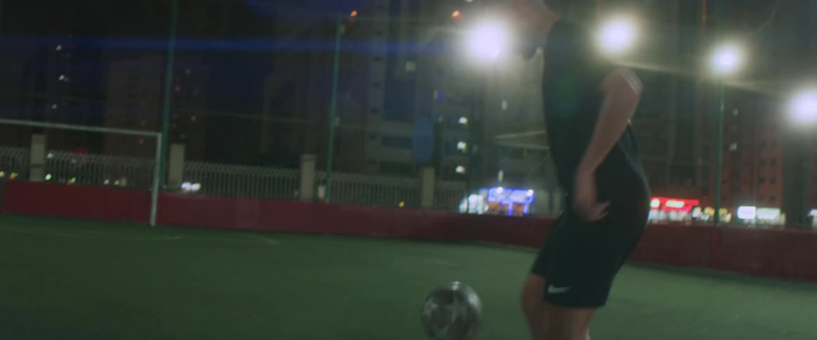 Male athlete practices with soccer ball on soccer field with skyscrapers in the background at night