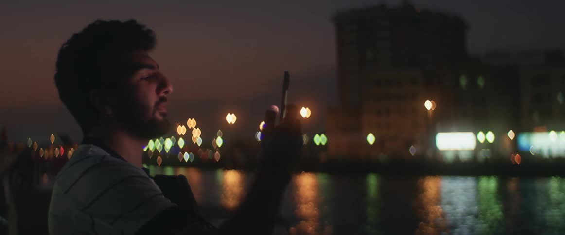 A man taking pictures on a smartphone in front of a city river at night
