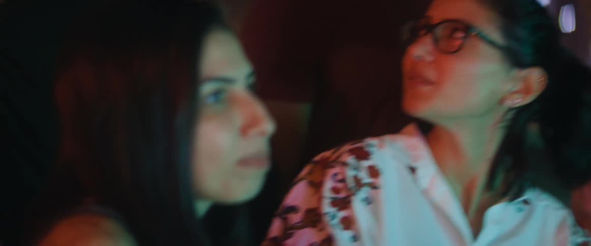 A group of men and women in a room watch something and then one woman turns and speaks to a woman behind her