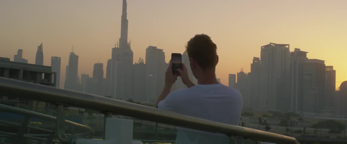 A man uses his smartphone to take a photograph of a city skyline at sunset