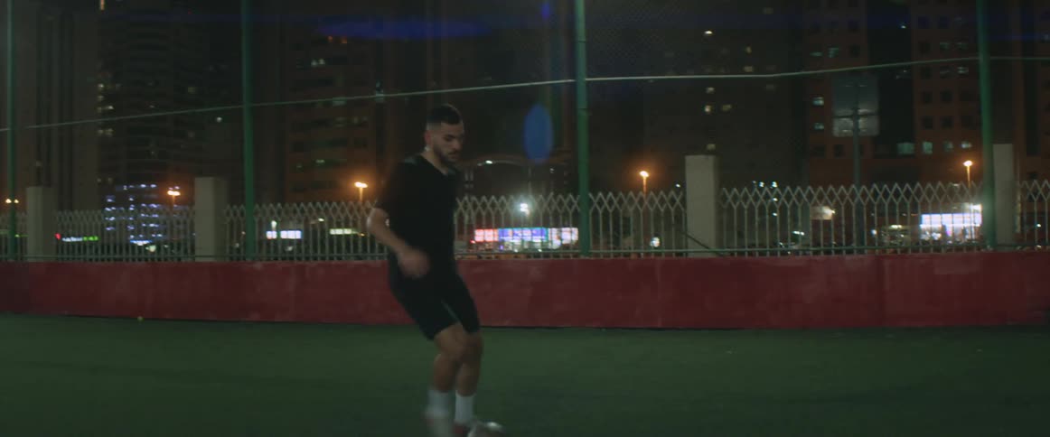 Athlete practices soccer footwork on field surrounded by buildings at night