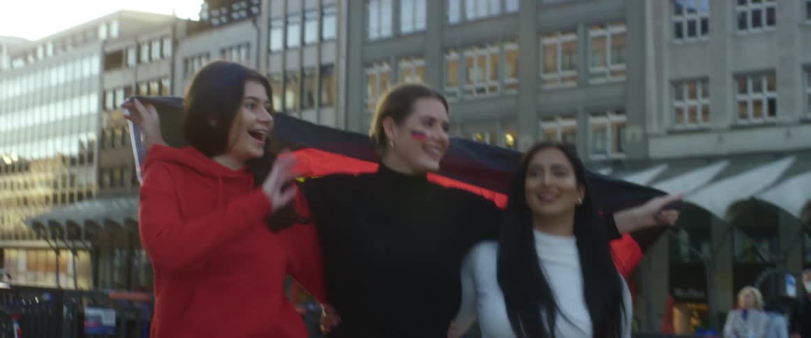 A trio of women walk together through a metropolis carrying the german flag over their shoulders