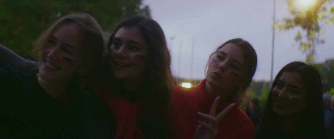 A group of young women pose with a german flag as they take a selfie with a smartphone outdoors in the evening