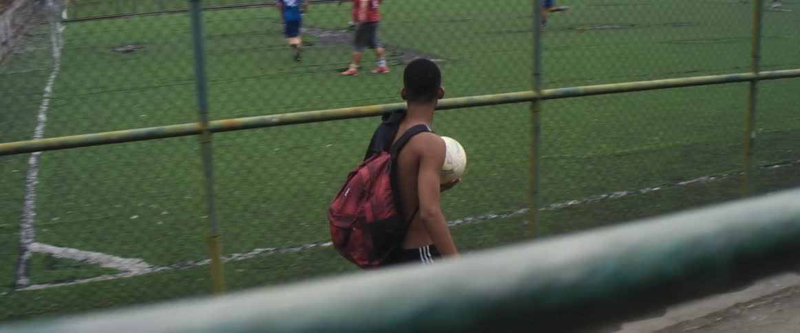 A teenager carrying a soccer ball and other boys walk towards a soccer field with people playing soccer
