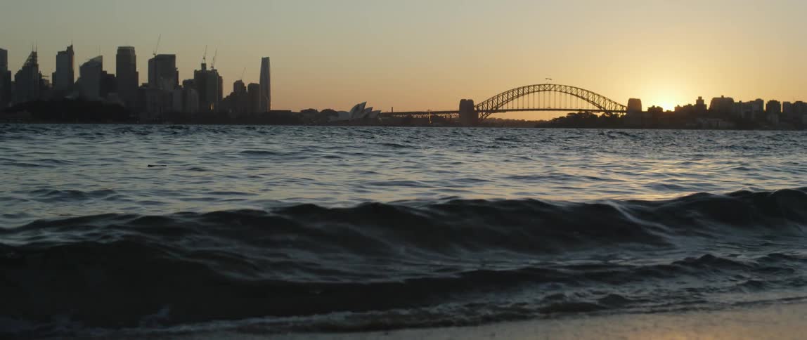 Waves are rolling on to a beach with tall city buildings and a bridge in the distance and the sun on the horizon