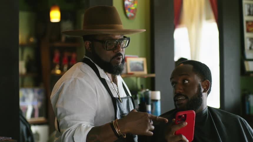 A man shows his barber something on his smartphone as he gets a haircut
