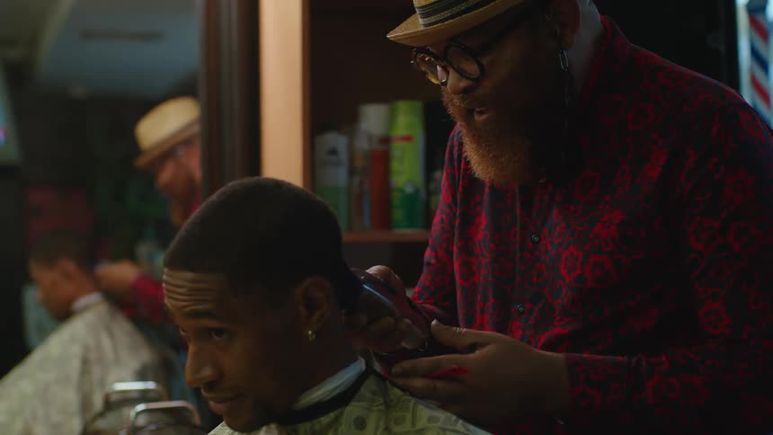 Barber with hat glasses red beard and red shirt cuts mans hair with clippers in barber shop