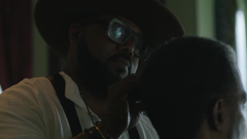 Barber with hat glasses beard white shirt and black apron trims mans hair with clipper in barber shop