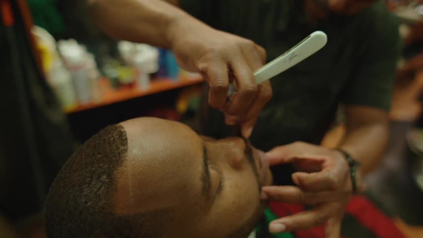 A male barber uses a straight blade to shave another mans beard