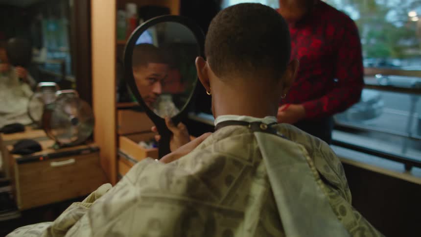 A man in a barber examines his face and hair in a mirror and nods as man stands by and watches him