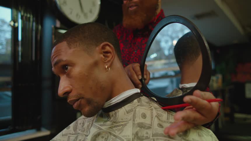 A barber holds a mirror behind the head of a customer so that the customer can examine their haircut in the mirror in front of them