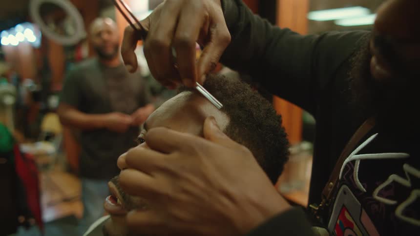 Barber uses straight razor on near clients forehead as a fellow worker looks on