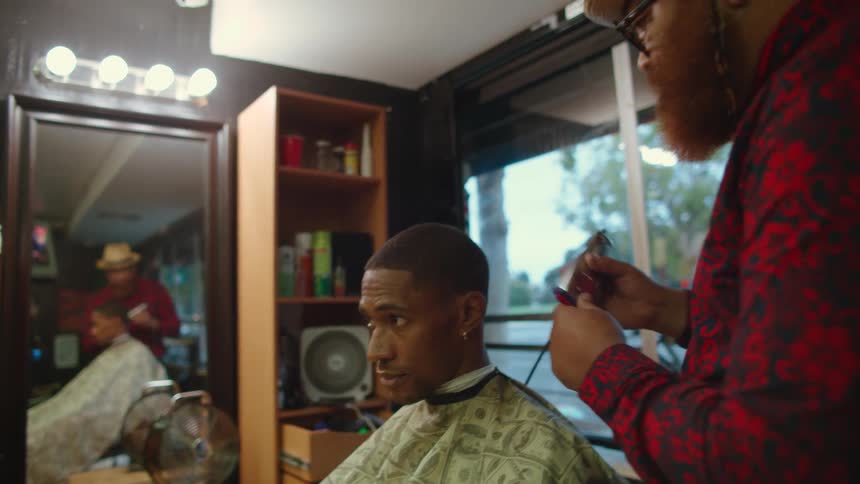 Barber with brown hat glasses red beard and shirt cuts mans hair with clippers in barbers shop
