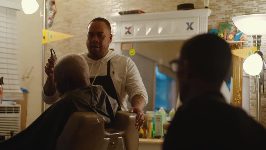 A barber shaves a mans head as another man sits and watches