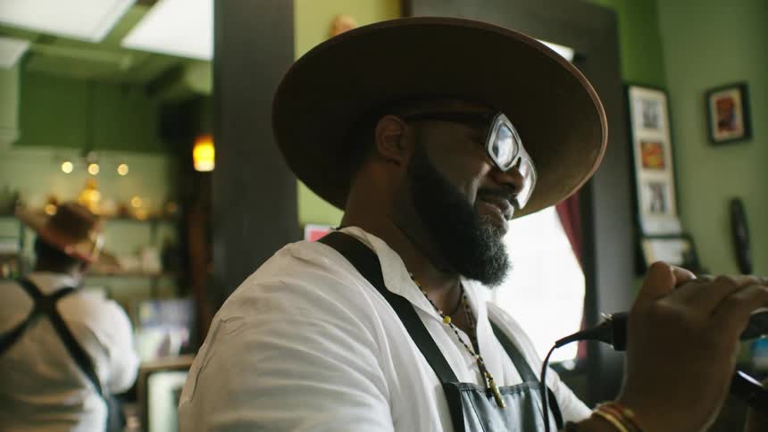 Barber with hat glasses beard white shirt and black apron talks with customer while cutting his hair with clippers smiling in barber shop