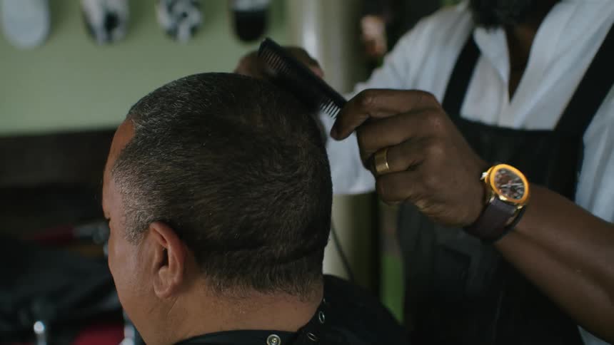 A barber uses a comb and hair trimmers to cut the hair of a customer in a barber shop during the day