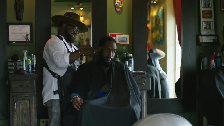 Barber with hat glasses beard white shirt and black apron cuts mans hair with clippers in barber shop talking and laughing