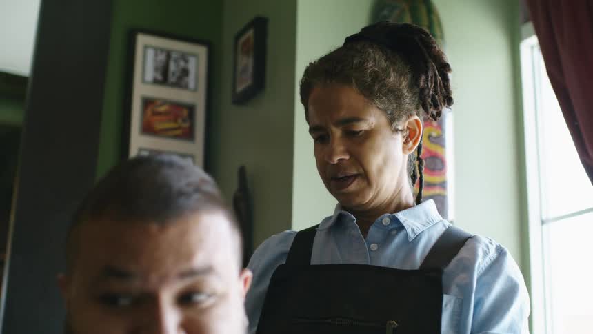 A man is sitting indoors during the day while a female barber stands behind him trimming his hair