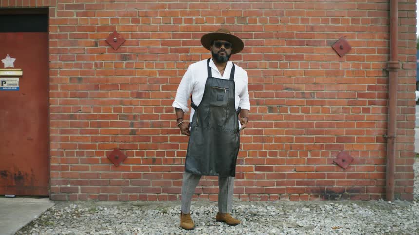 A man wearing a work apron walks in front of the camera and poses before a brick wall