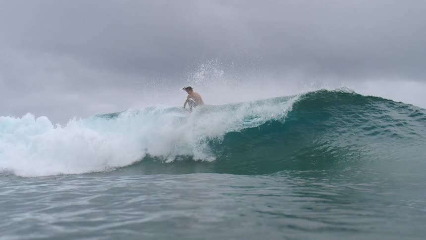 A surfer wearing shorts rides a big wave under a cloudy sky