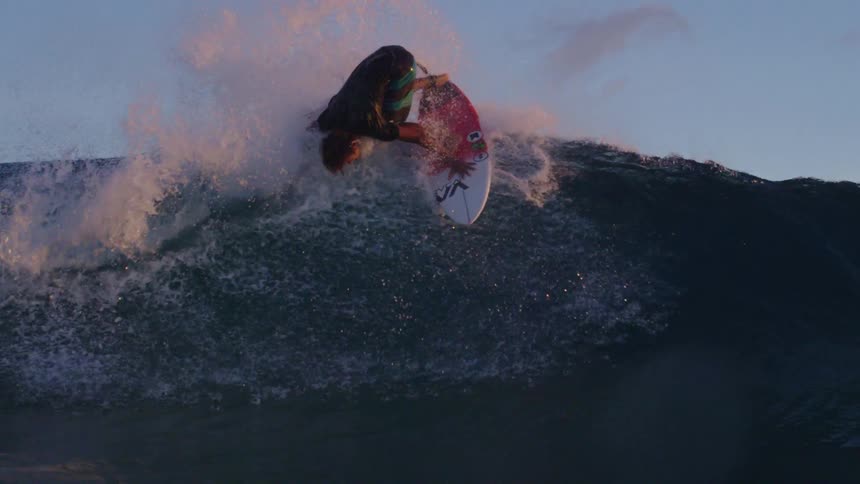 A surfer wearing shorts and a dark colored t shirt rides a big wave