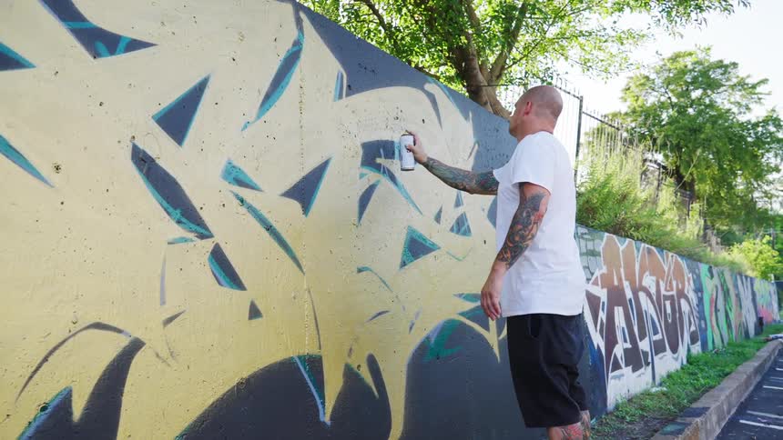 Young bald tattooed man spraying graffiti on a wall in the parking lot