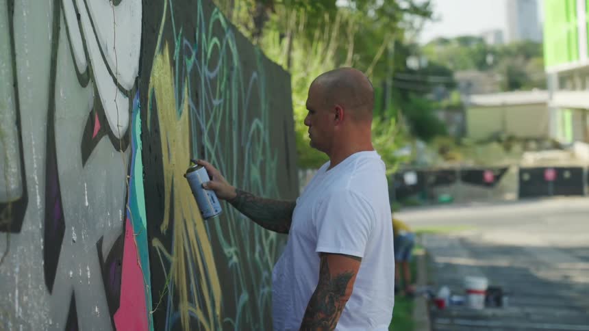 A man is spray painting a wall outdoors with graffiti